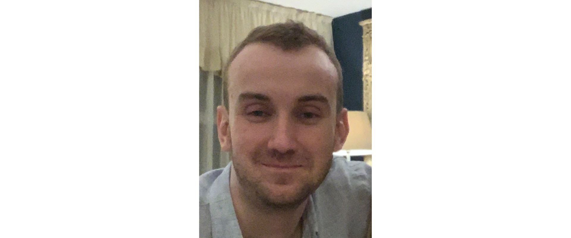 Portrait of a person in a light blue shirt, photographed indoors with curtains and a lamp in the background.