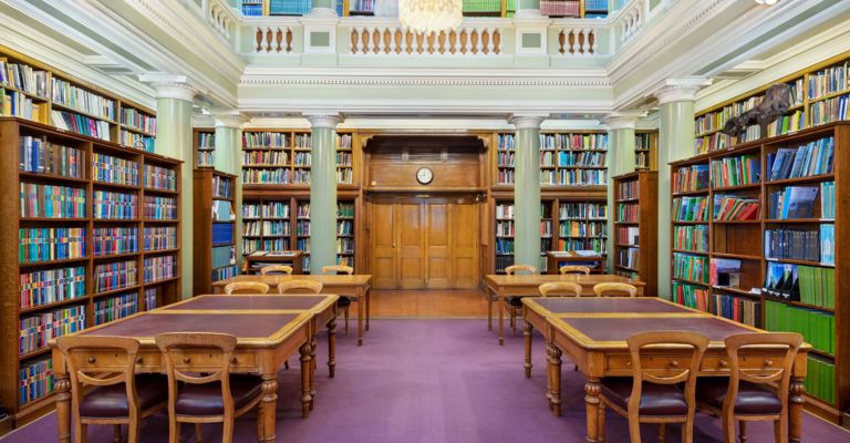 The Upper Library of the Geological Society of London, tables and chairs on a maroon carpet surrounded by book-lined shelves and green pillars