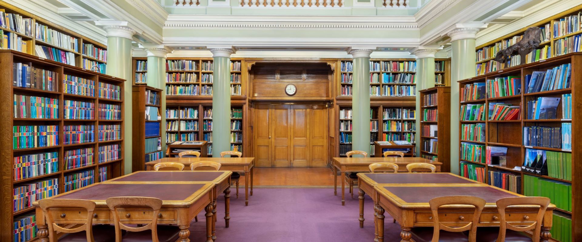 The Upper Library of the Geological Society of London, tables and chairs on a maroon carpet surrounded by book-lined shelves and green pillars