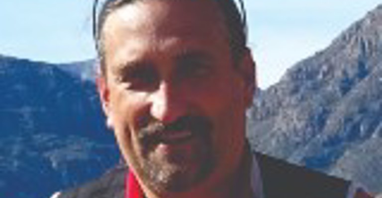 A man with a short beard with sunglasses on his head, wearing a red fleece standing before a mountainous background