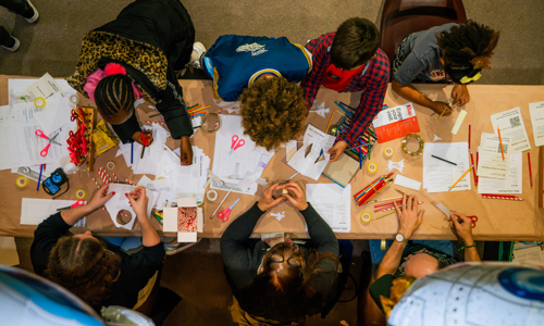 Looking down on a group of children sitting at a rectangular table working on activities with paper, scissors and crayons