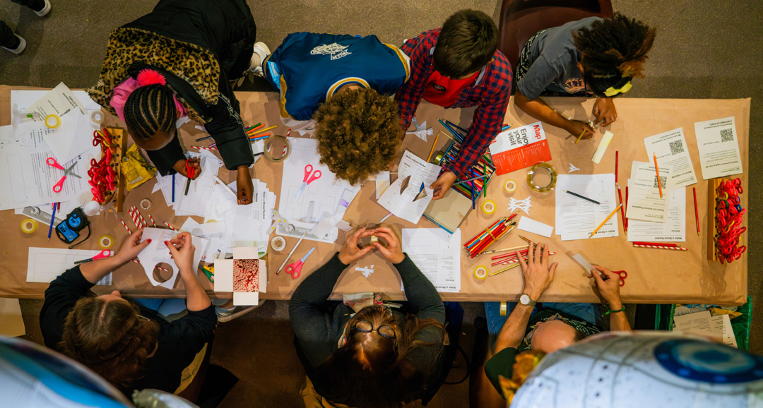 Looking down on a group of children sitting at a rectangular table working on activities with paper, scissors and crayons