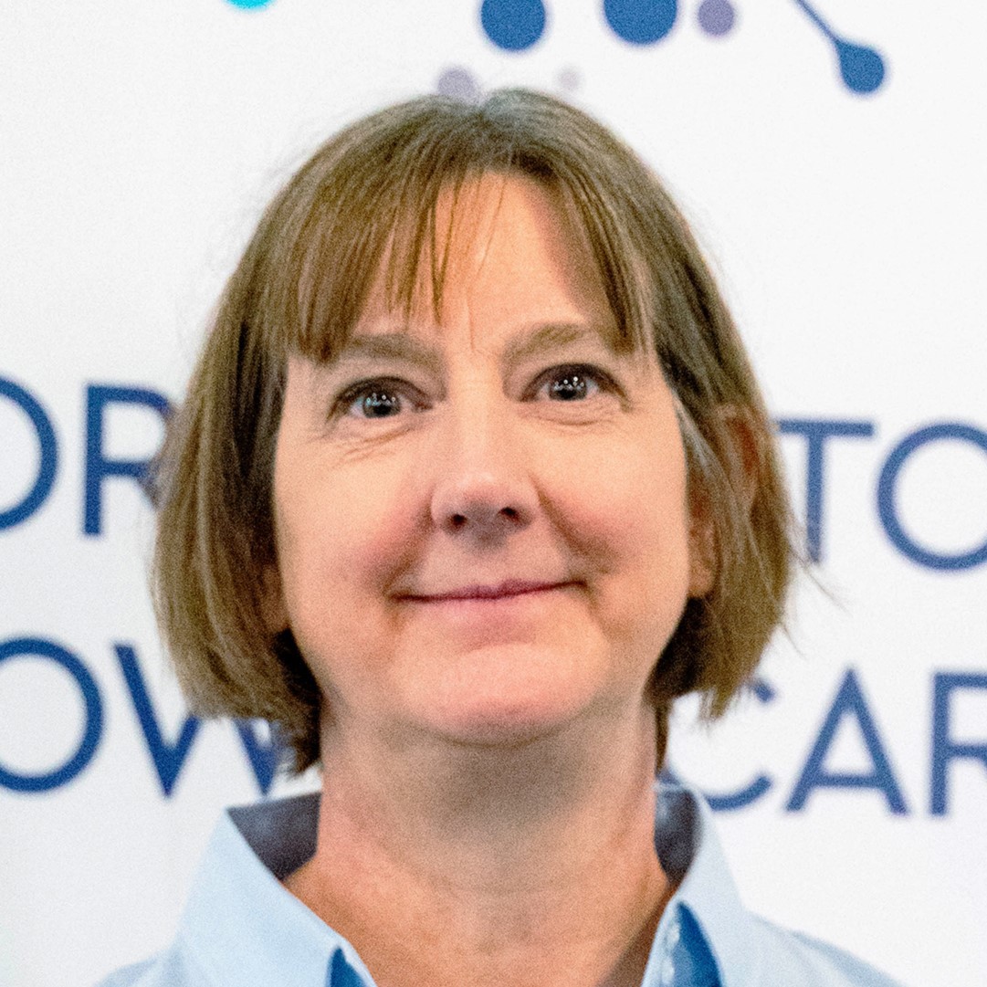 A person with short hair stands in front of a backdrop featuring blue text on a white background, conveying a professional setting.