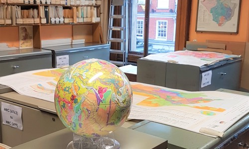 The Map Room of the Geological Society. A Room with orange walls, grey metal map cabinets, boxes of maps on shelves against the walls. Some geological map sheets are laid on the cabinets, as well as a geological map globe of the earth