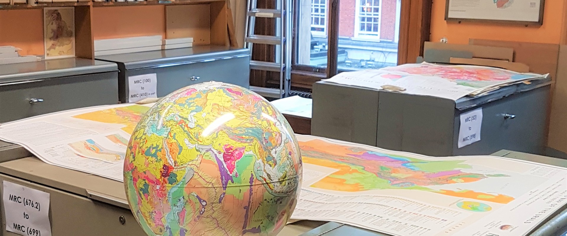 The Map Room of the Geological Society. A Room with orange walls, grey metal map cabinets, boxes of maps on shelves against the walls. Some geological map sheets are laid on the cabinets, as well as a geological map globe of the earth