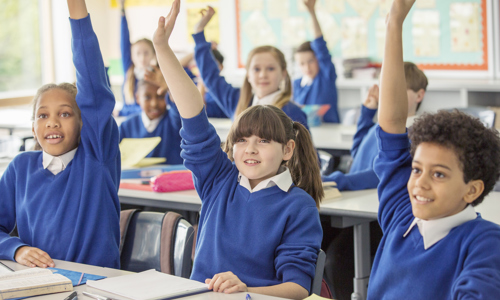 primary school age children of different ethnicities and genders with their hands up in the classroom