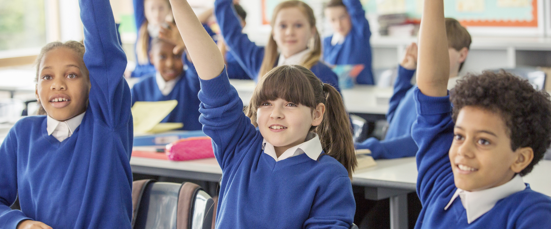 primary school age children of different ethnicities and genders with their hands up in the classroom