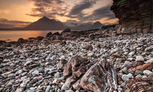 A view of a pebbly beach in the foreground, looking cross a stretch of water towards Cuillin Ridge mountain in Scotland, with the sun setting behind it
