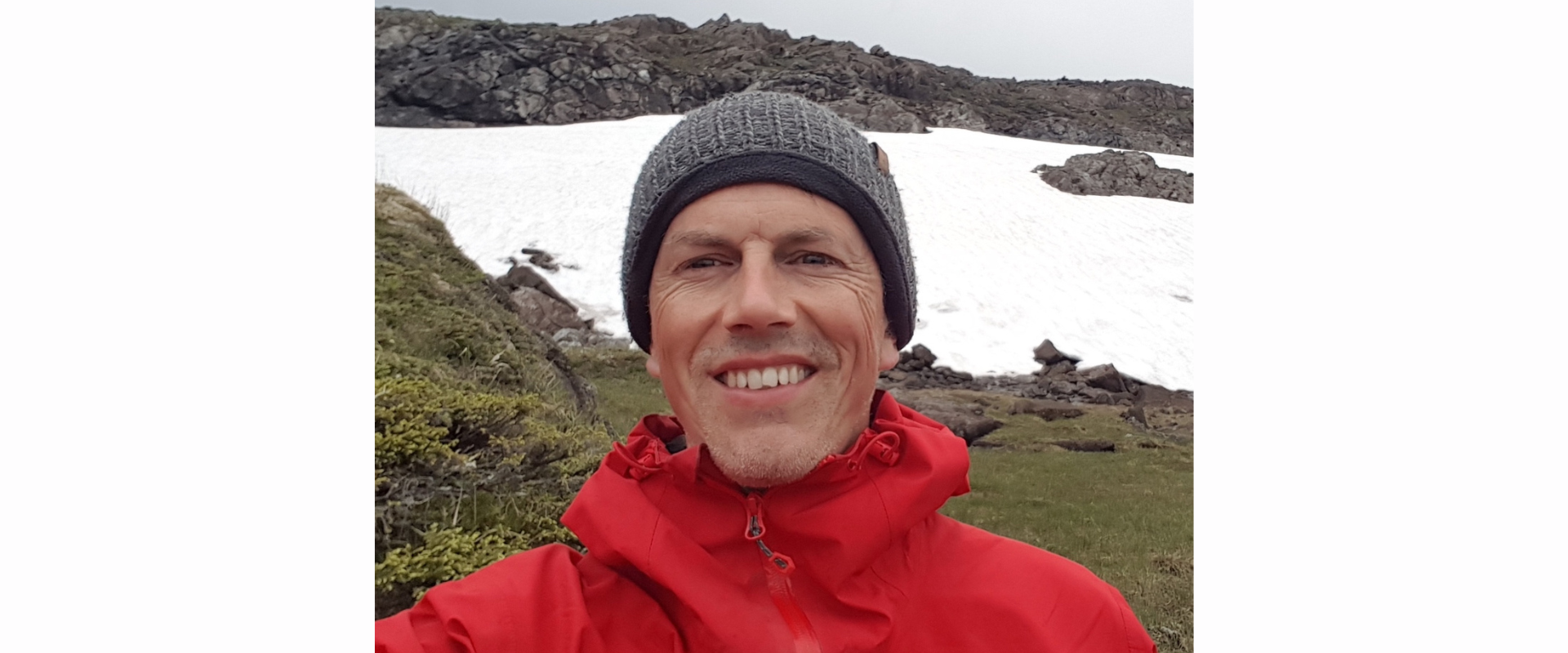 A person wearing a bright red outdoor jacket and a knitted grey hat, standing in a mountainous area with patches of snow on rocky slopes and green grass in the foreground.