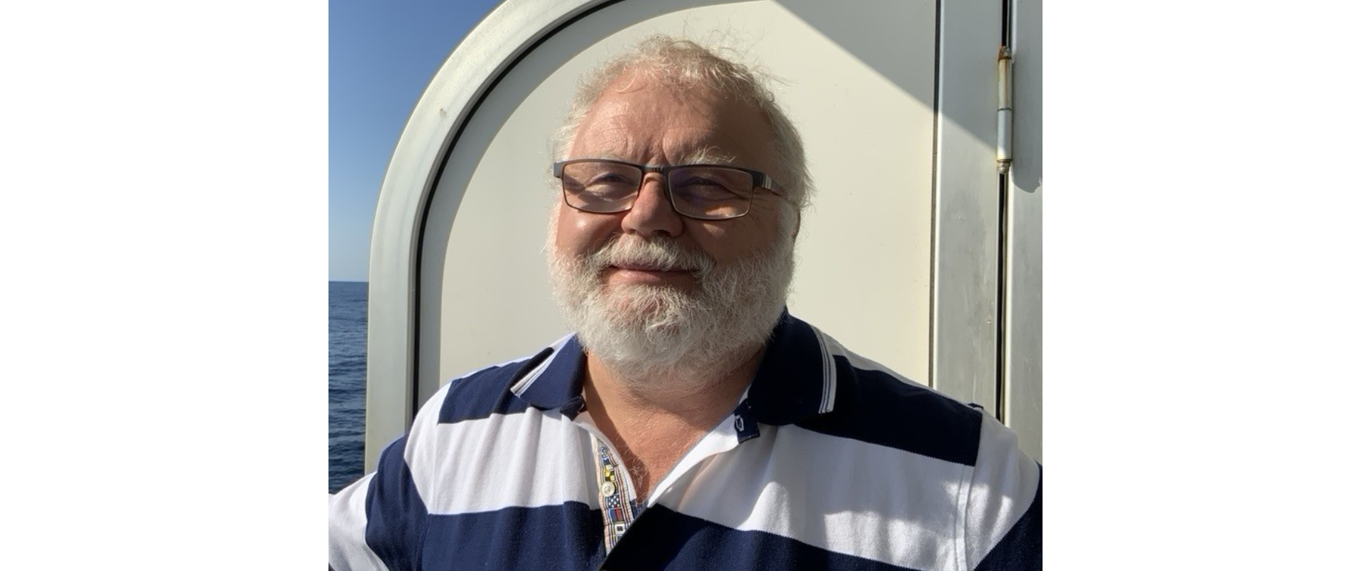 A person with white hair, a full white beard, and glasses stands outdoors on a vessel or platform near the sea. The person is wearing a navy-and-white striped collared shirt. Behind the person is a light-colored curved metal structure, with calm blue water and a clear sky visible in the background. Bright sunlight illuminates one side of the scene, creating strong shadows on the structure.