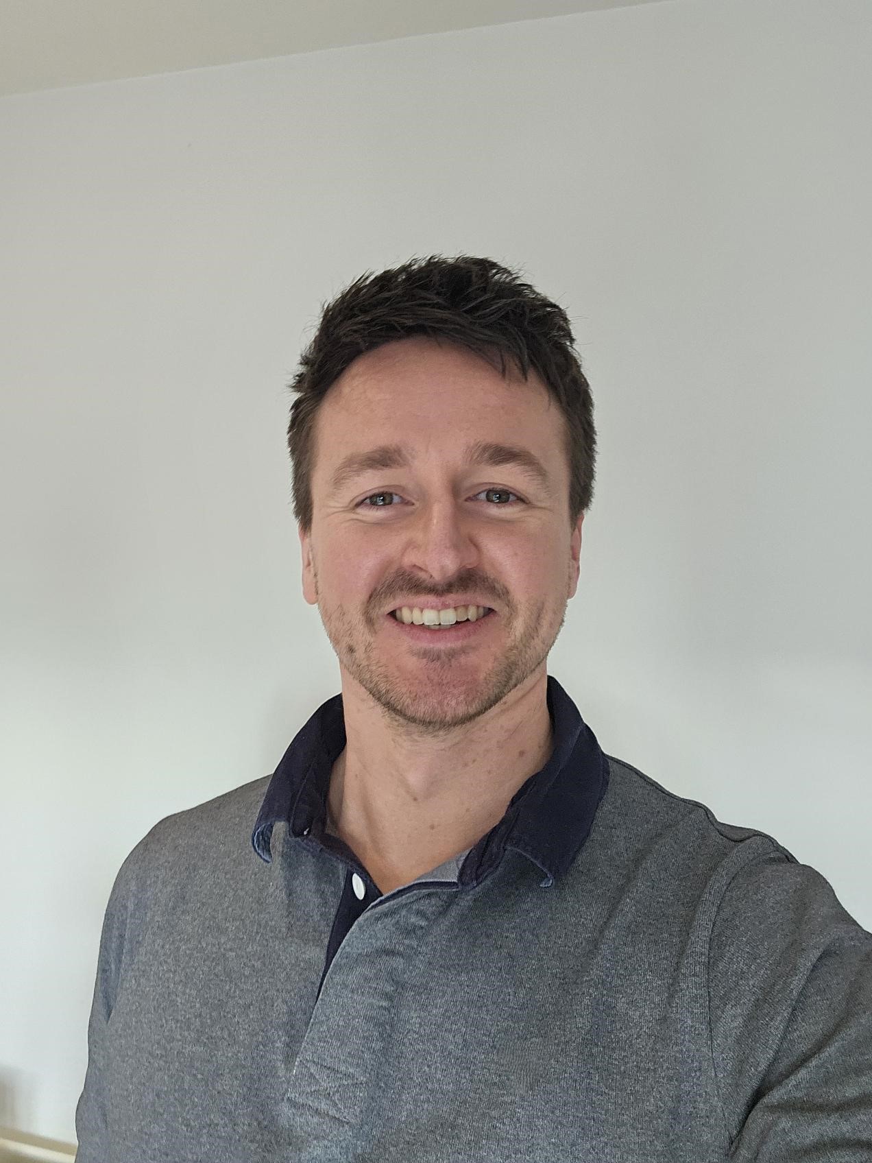 A person with short dark hair is facing the camera, wearing a collar shirt against a plain background.