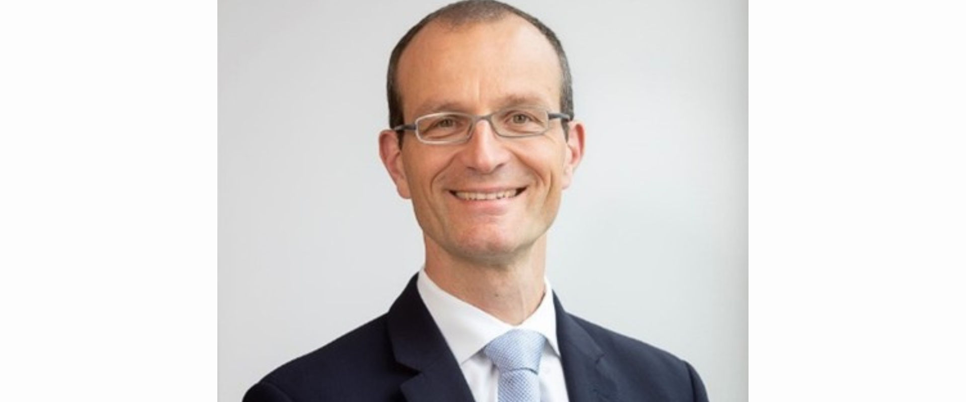 A person wearing a dark suit jacket, a white shirt, and a light‑coloured tie, posed against a plain light background.