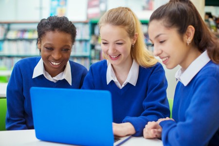 Two white girls and one black girl all in school uniform of blue jumpers and white blouses, all smiling looking at a laptop