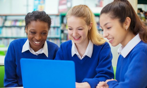 Two white girls and one black girl all in school uniform of blue jumpers and white blouses, all smiling looking at a laptop