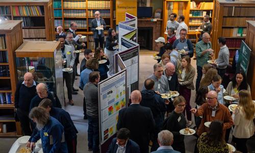 A lively networking event in a library, with attendees enjoying food and discussing onlookers among colorful posters and bookshelves.