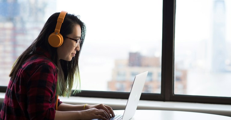 A woman with long hair wearing a red check shirt and orange headphones sitting at a laptop in front of a window