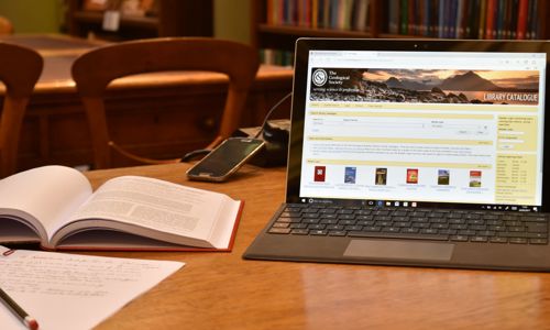 A table in a library with a laptop showing a library catalogue, a mobile phone, an opened book and a page of writing with a pencil. Bookshelves in the background.