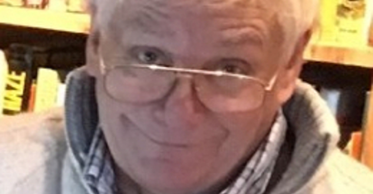 A man with white hair wearing glasses in front of shelves of books, looking at the camera