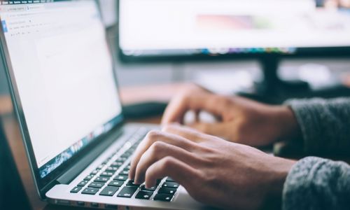 A laptop with two hands typing on it, with a PC blurred in the background