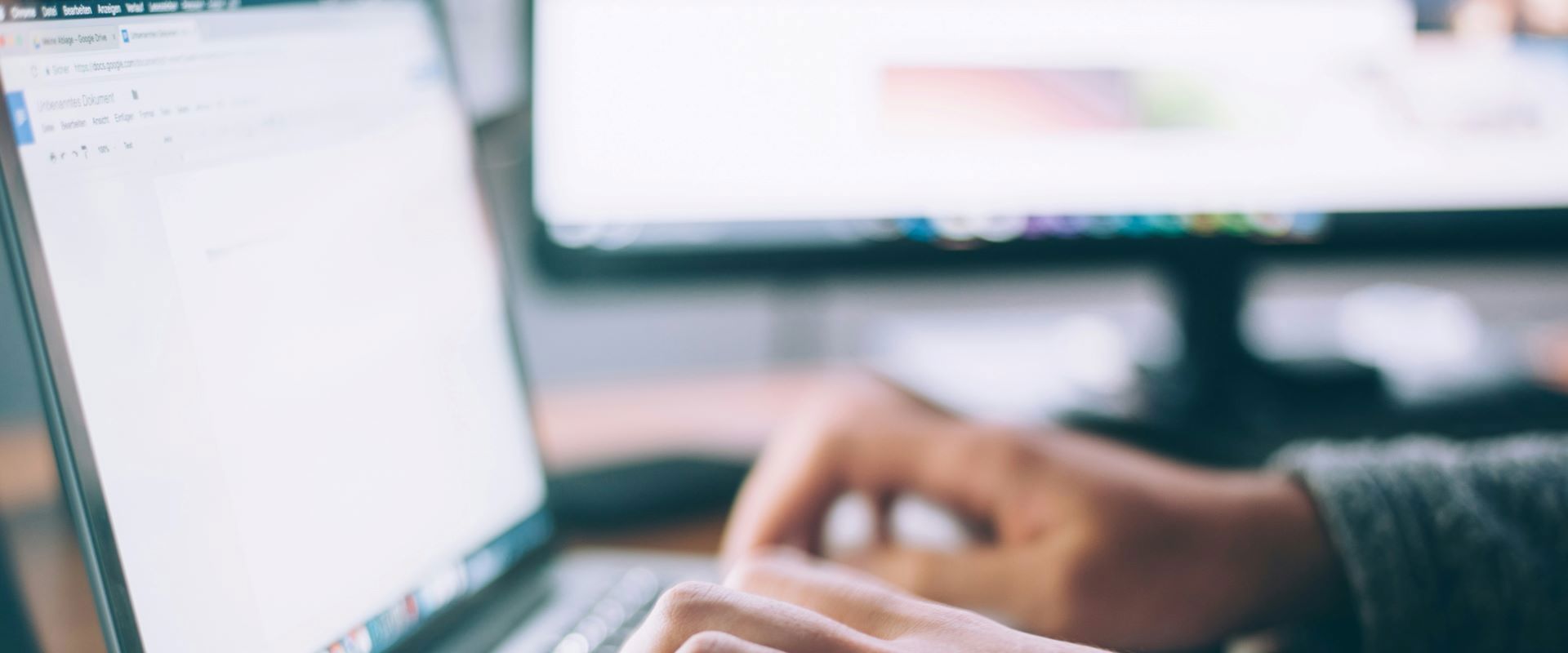 A laptop with two hands typing on it, with a PC blurred in the background