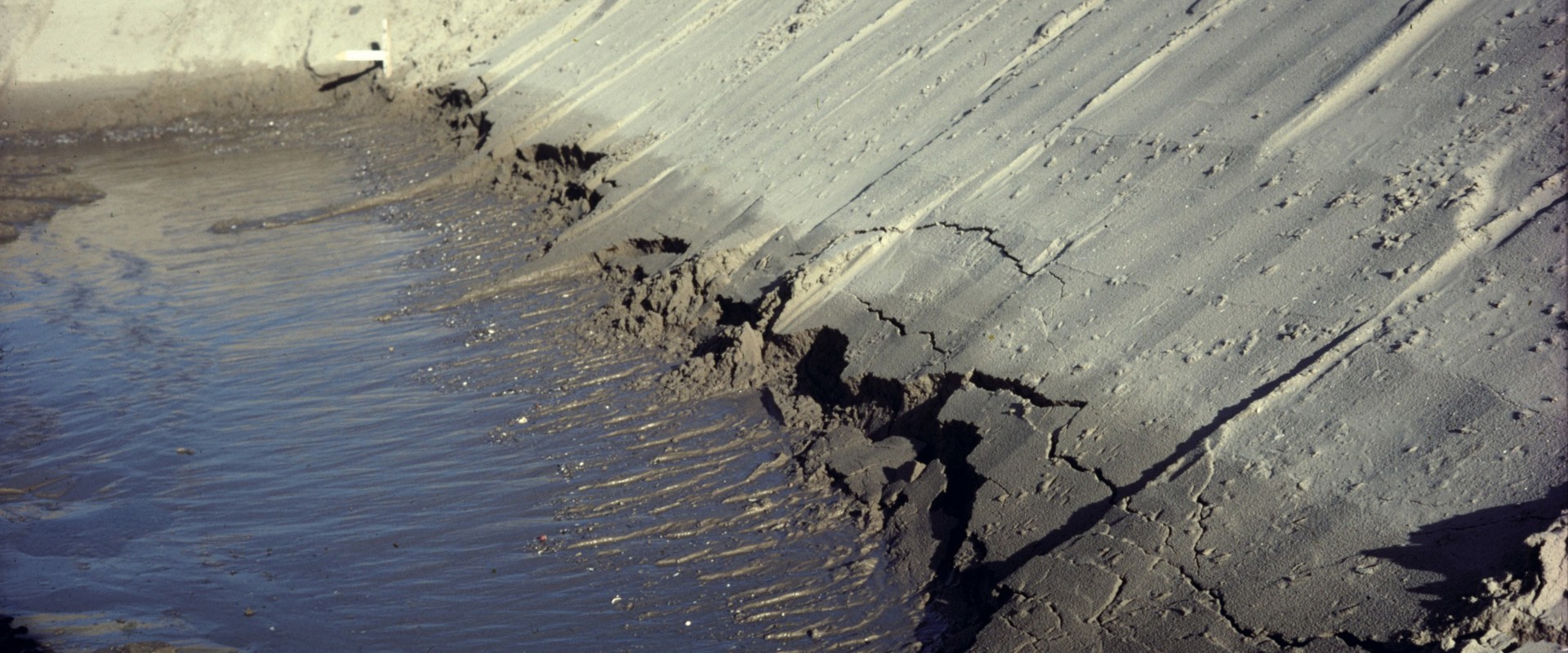 "Erosion of a sandy embankment beside a body of water, showing cracks and partially collapsed soil near the water's edge."