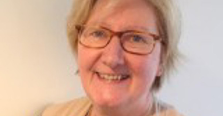 A woman with short fair fair wearing glasses and a sleeveless beige cardigan over a black top, smiling at the camera