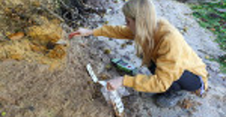 A researcher in a yellow jacket examines soil layers at an excavation site, brushing away dirt with a tool.