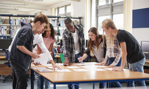 seven young people of differing ethnicities stood around a table smiling and looking at papers on the table