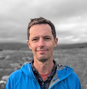 A person stands outdoors wearing a blue jacket over a plaid shirt, with a blurred background of gray skies and rocky terrain.