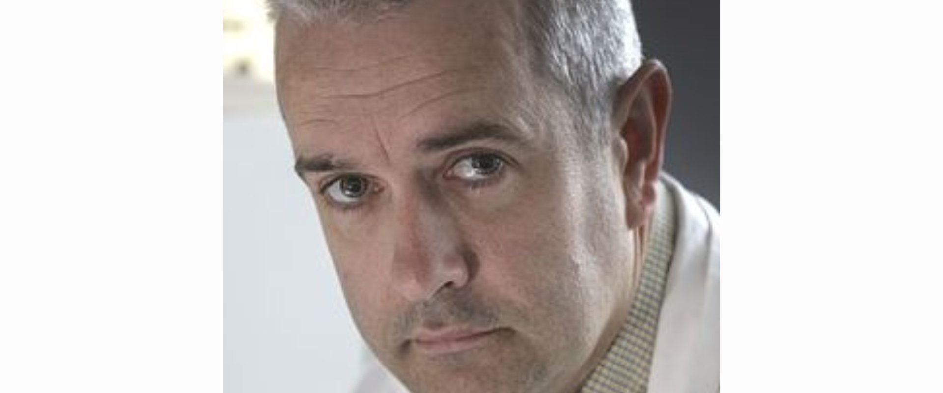 A person wearing a light‑coloured collared shirt with a white coat layered over it, positioned indoors against a softly lit background.