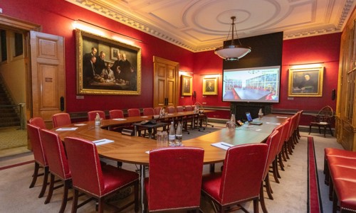 large boardroom table surrounded by red leather seating and red walls with period paintings adorning them, wood panelled doors and an ornate white ceiling