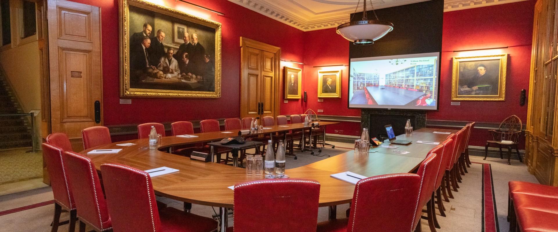 large boardroom table surrounded by red leather seating and red walls with period paintings adorning them, wood panelled doors and an ornate white ceiling