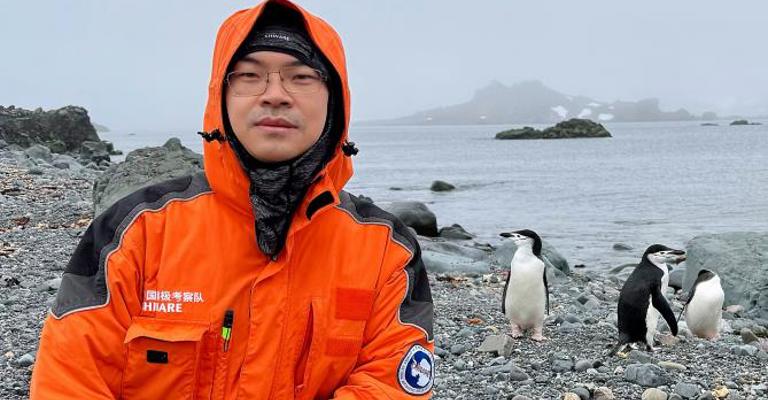 A man wearing glasses and an orange coat with the hood up, kneeling on a stony beach with penguins in the background