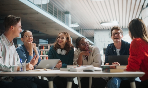 A group of young people around a table with laptops and textbooks laughing