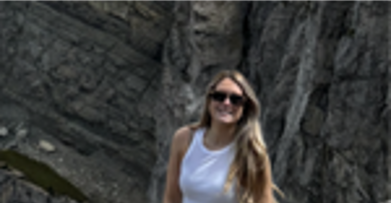 A woman with long blonde hair wearing a white top, white shorts and sunglasses standing in front of a rocky background and smiling