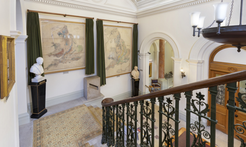 The staircase of the Geological Society of London, leading down stairs to the Society's entrance hall, with two large geological maps on the walls, and busts of George Greenough and William Smith