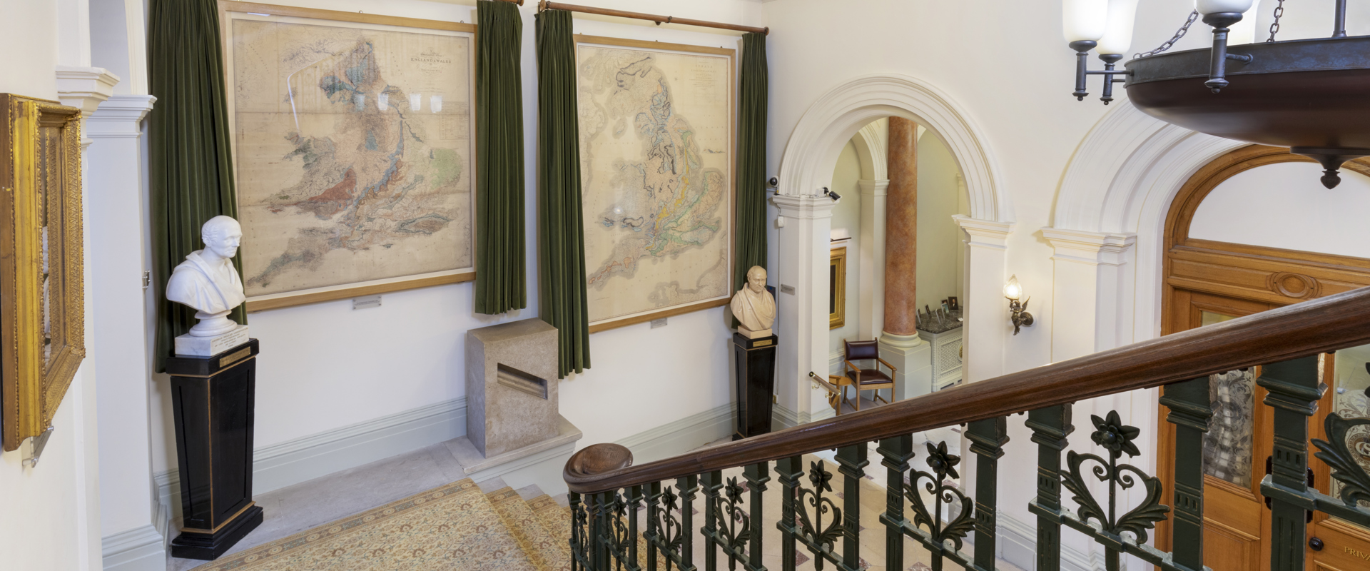 The staircase of the Geological Society of London, leading down stairs to the Society's entrance hall, with two large geological maps on the walls, and busts of George Greenough and William Smith