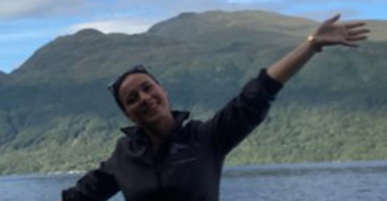 A woman sitting down in front of a lake and a mountain with her arms outstretched, smiling
