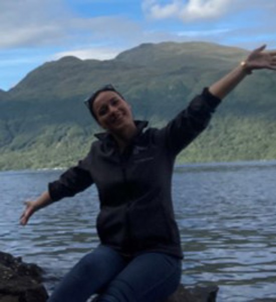 A woman sitting down in front of a lake and a mountain with her arms outstretched, smiling