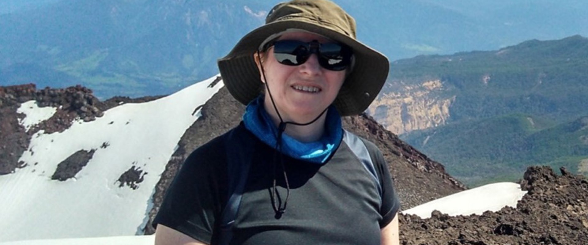 A smiling person wearing a hat and sunglasses stands on a rocky, snow-topped mountain with a distant volcano and vast landscape under a bright sky.