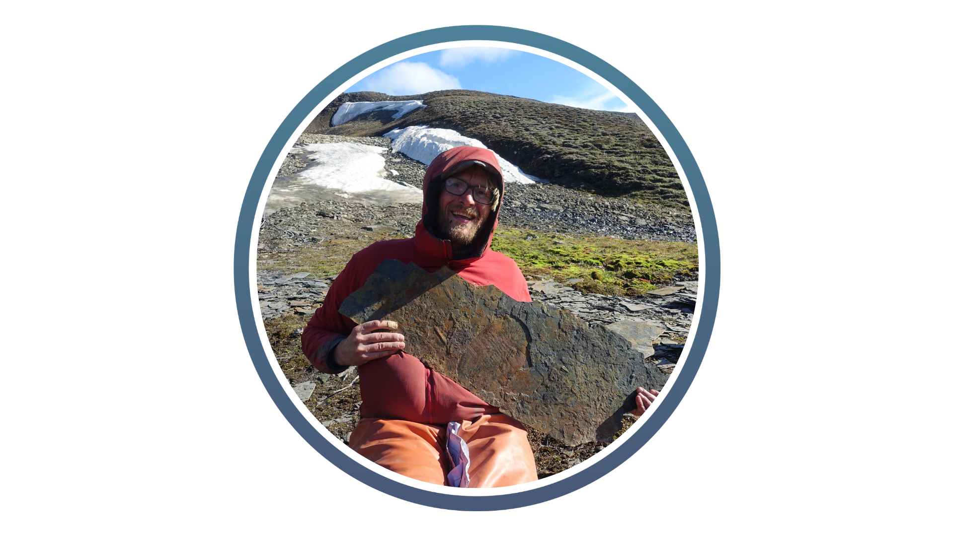 Person in outdoor gear holding a large rock slab with fossil imprint, set against a mountainous landscape with patches of snow.