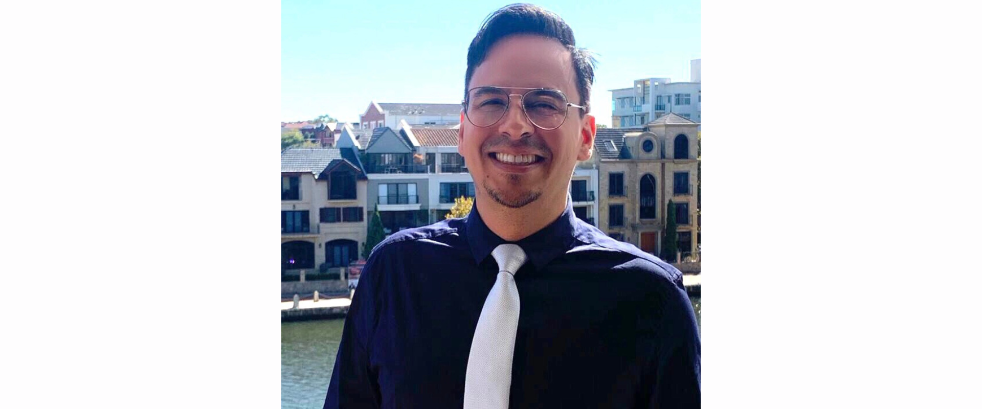A person wearing a dark long‑sleeved shirt with a white tie, standing outdoors in front of a waterfront area lined with multi‑storey buildings featuring balconies and varied architectural styles.