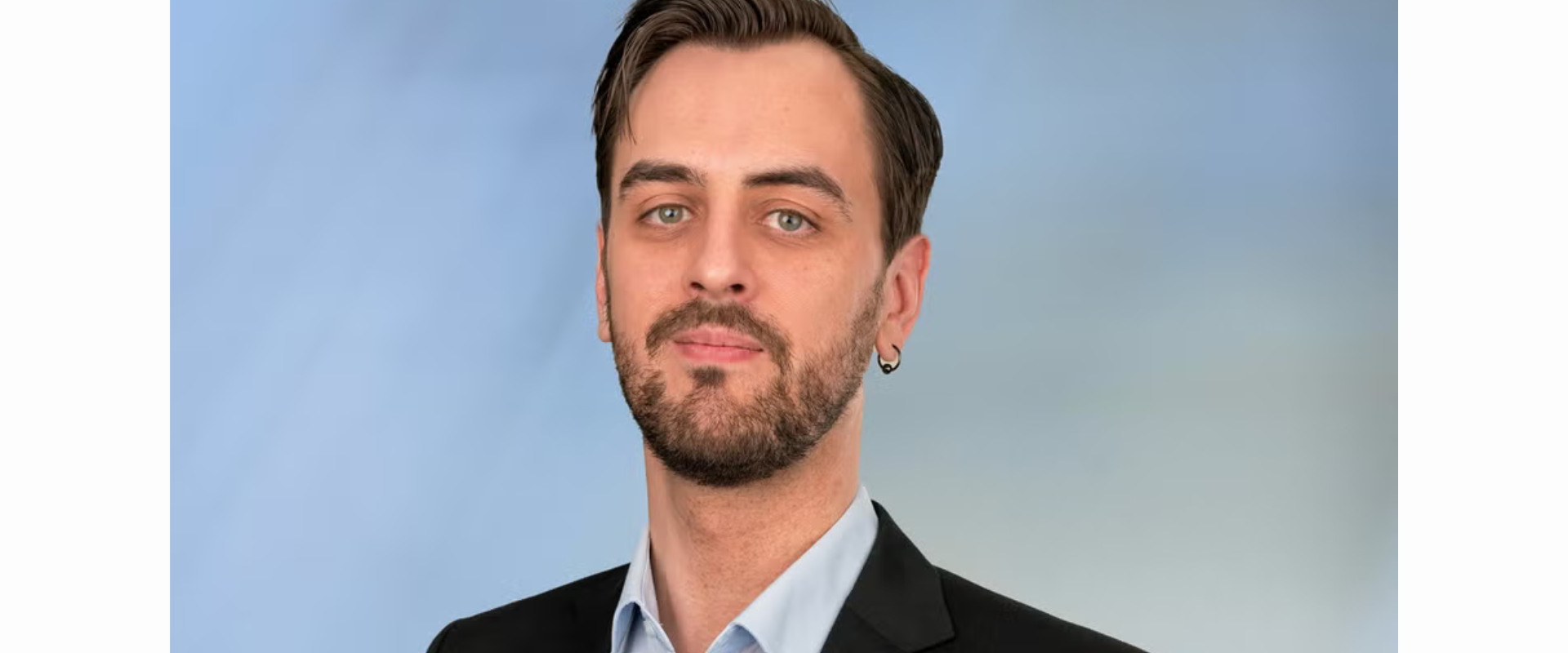 A person wearing a light blue collared shirt and a dark suit jacket, positioned against a soft blue gradient background. The person has short, neatly styled hair and is shown from the shoulders up.