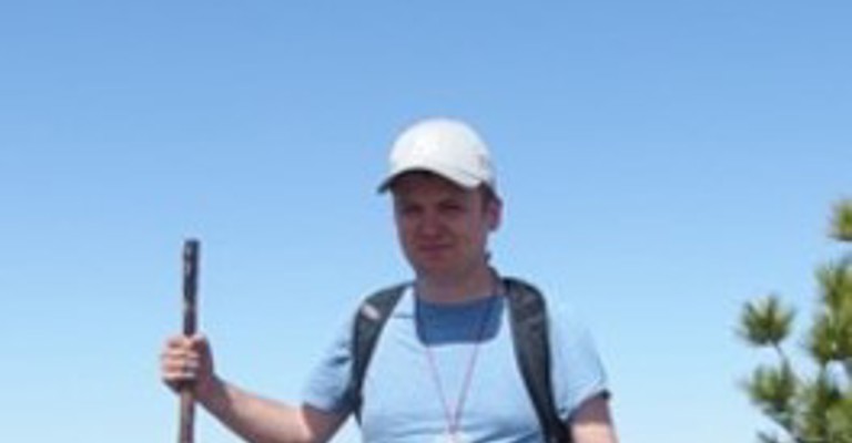 A man wearing a pale blue teeshite, dark grey trousers, white cap and a backpack holding a pole standing in front a mountainous landscape