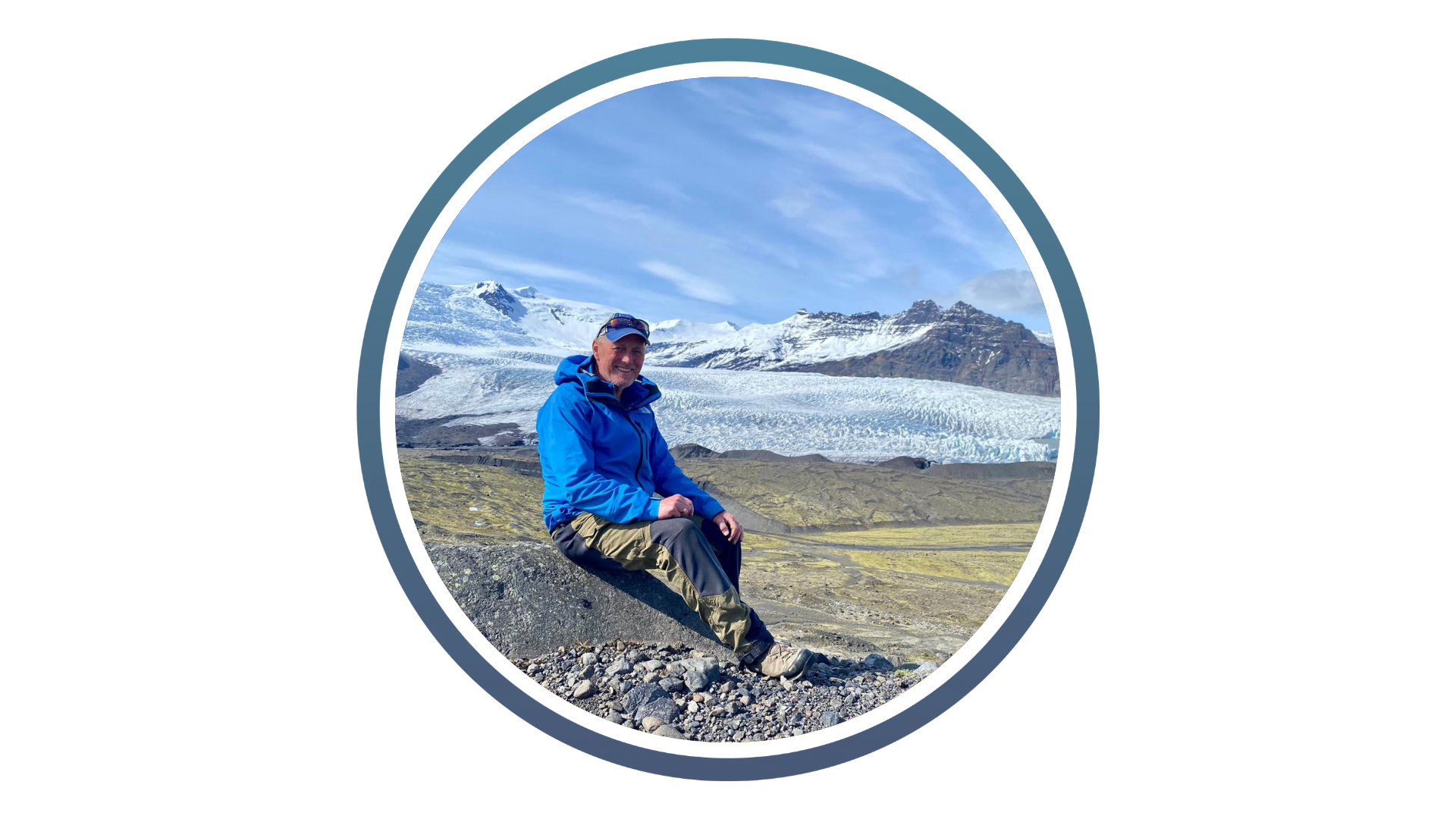 man in blue jacket with a cap on sat in front of a snow capped mountain range